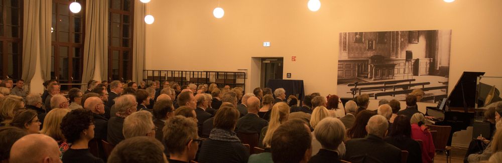 Ausschnitt aus dem Emil Berliner Saal in der ehemaligen Samsonschule. Zu sehen ist die Eröffnungsveranstaltung in Reihenbestuhlung mit dem Flügel. Im Hintergrund eine vergrößerte historische Fotografie der führeren Aula.